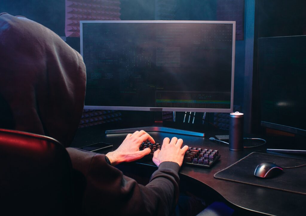 homem de capuz preto digitando de frente a um computador com códigos na tela